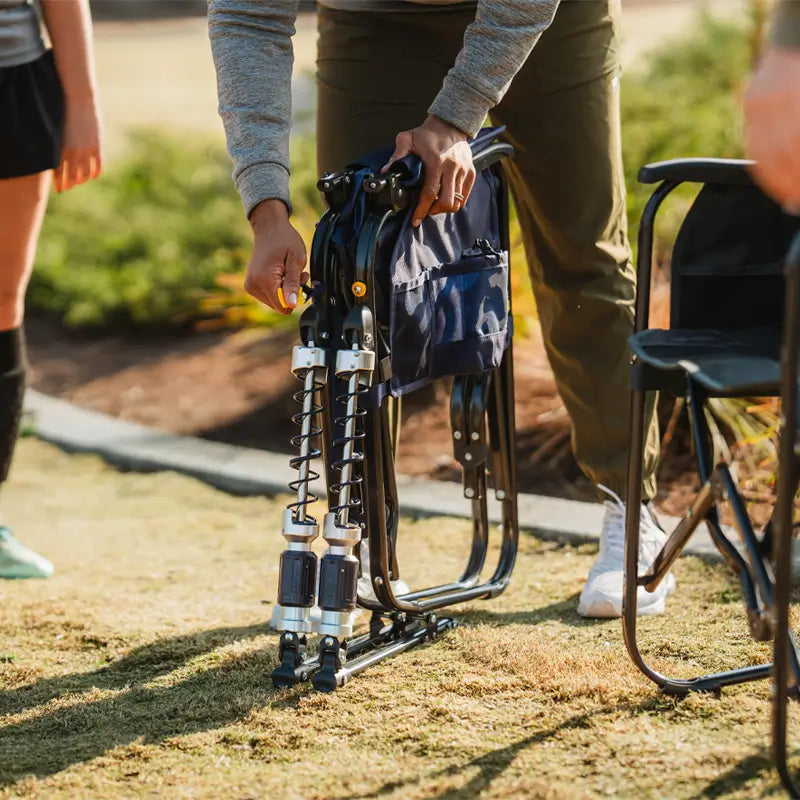 Person prepares to set up a folded navy Adjustable Rocker chair on a grassy lawn.