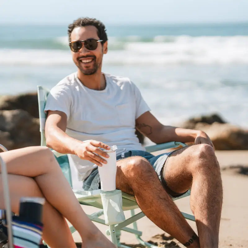 Man relaxing in the Green Beach Wave beach rocker with a drink in hand, seated on a sunny beach.