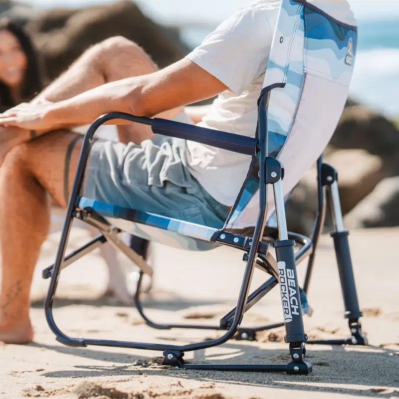 Man sitting on a Blue Beach Wave beach rocker with white frame, relaxing on the beach in front of ocean rocks.