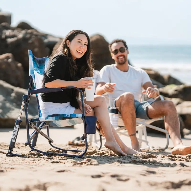 Woman smiles while holding a drink on a Blue Beach Wave beach rocker, chatting with a man beside her on the beach.