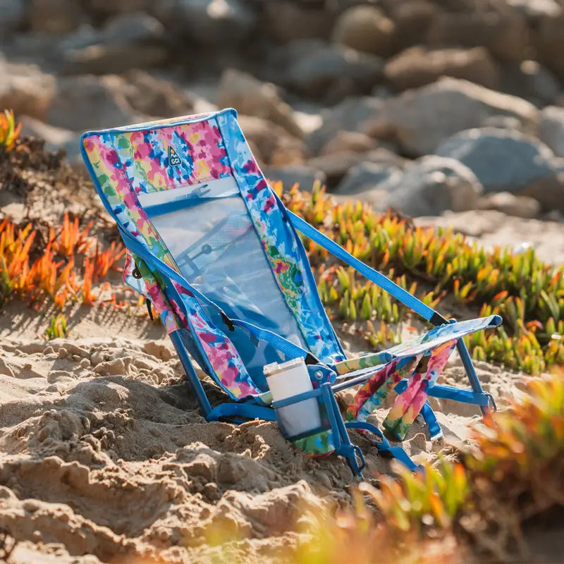Tie dye Everywhere Chair 2 set up on sand near beach plants with drink in cupholder.