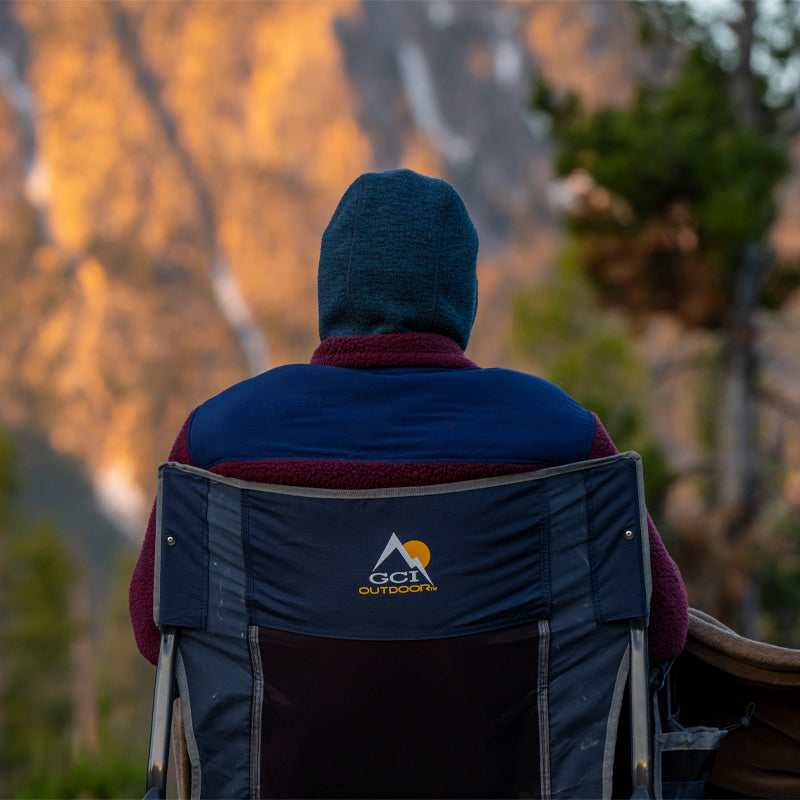 Person in a hooded jacket sits in an indigo Freestyle Rocker XL chair, gazing at a sunlit mountain.