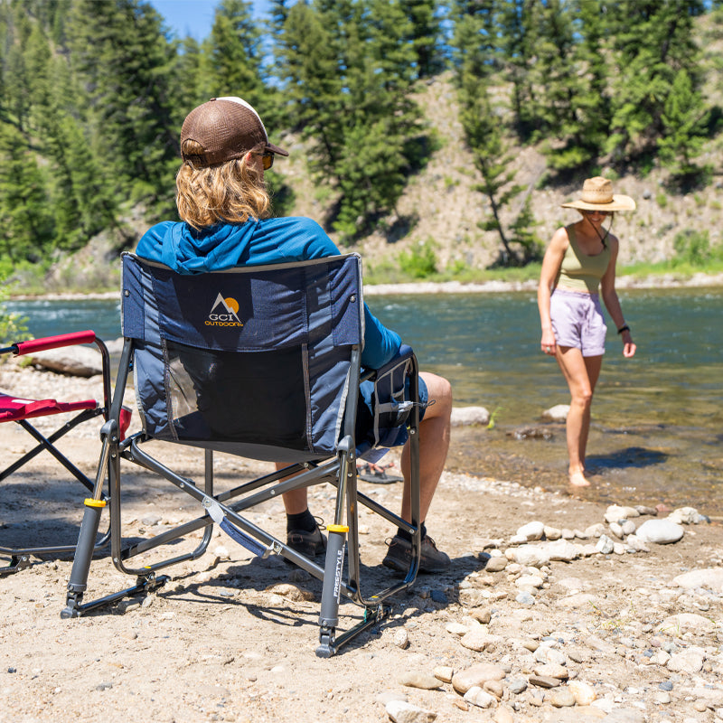 Man relaxes in an indigo Freestyle Rocker XL chair by a river while a woman walks near the water.