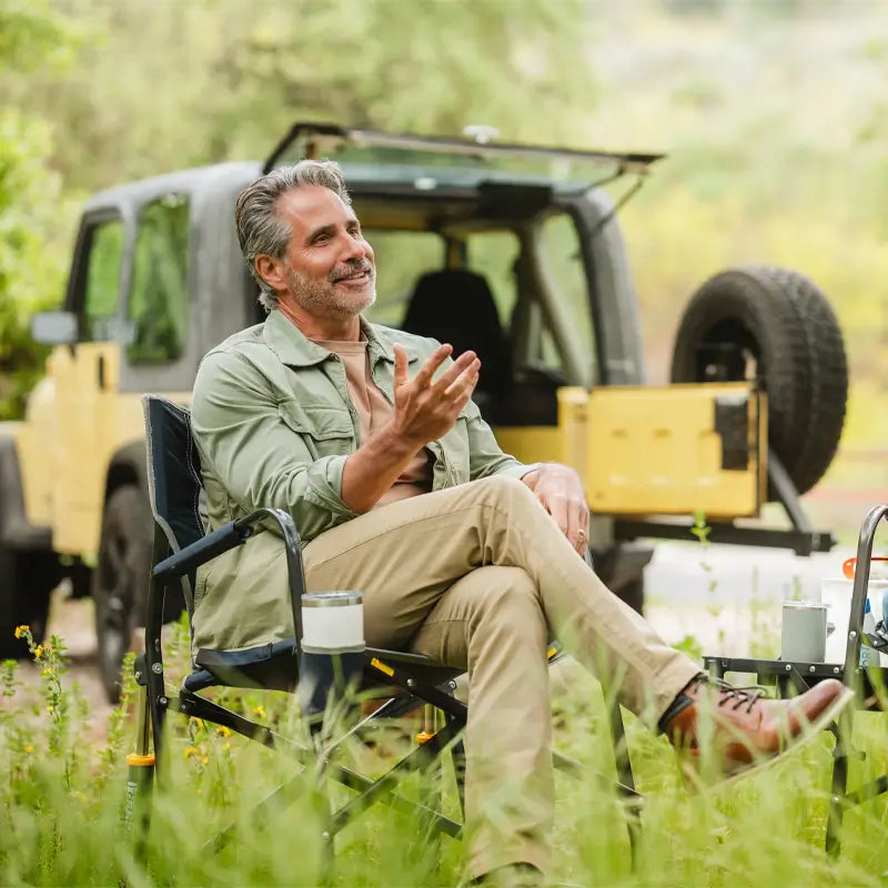 Man relaxing in a Freestyle Rocker near a parked Jeep in a wooded campsite.