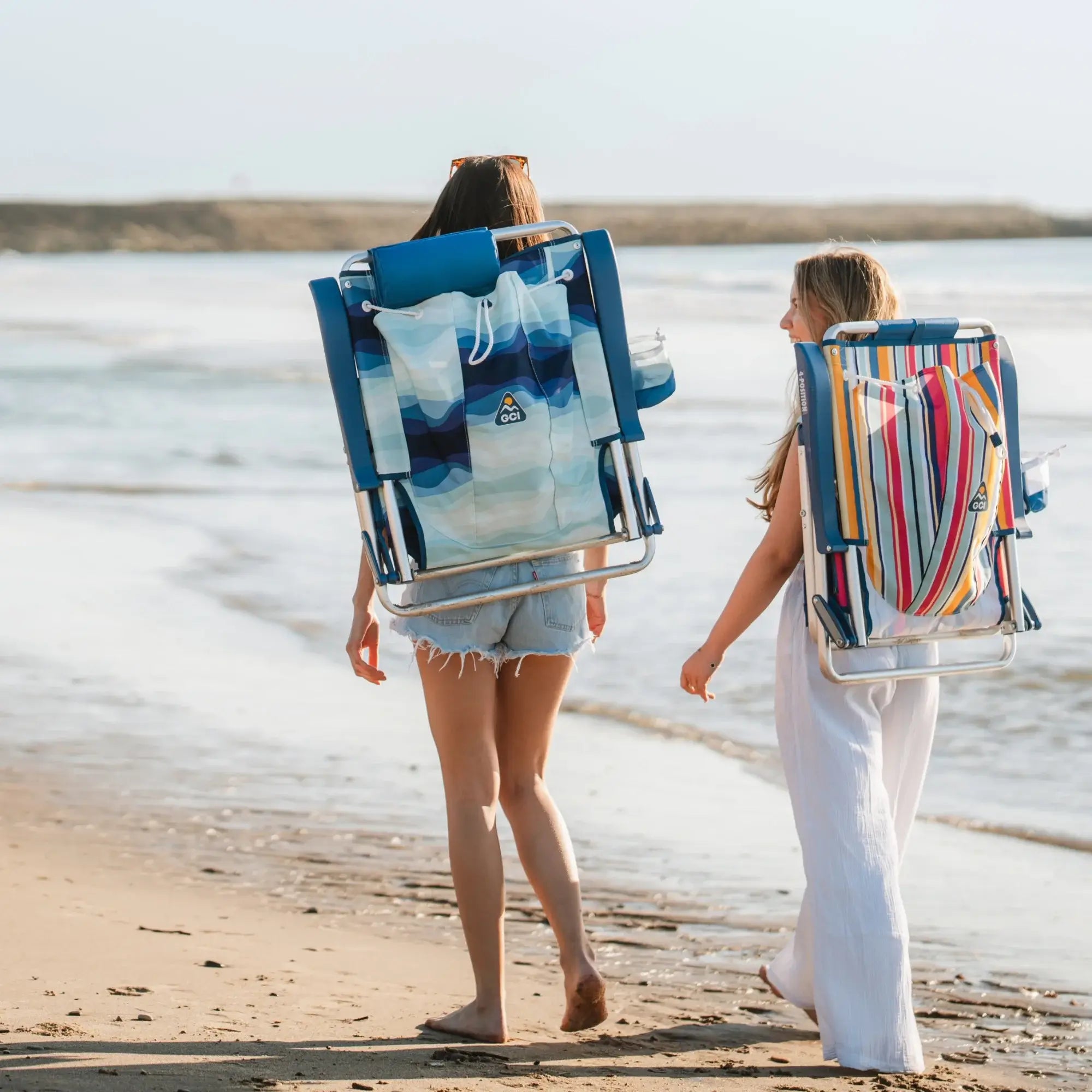 Two women walking on the beach with their backpack beach chairs attached to their back.