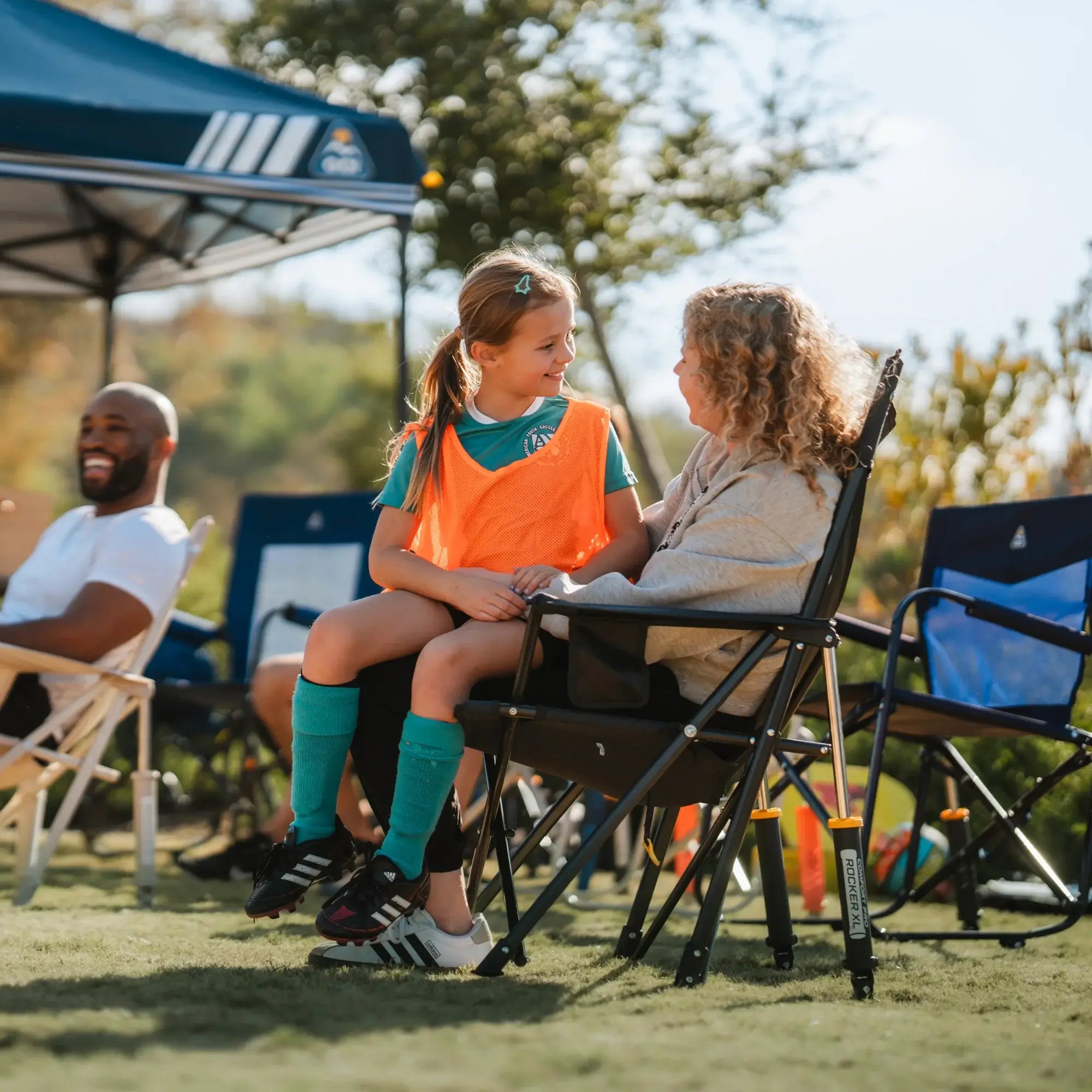 Two young girls sit together in a Comfort Pro Rocker XL at a soccer game, smiling and chatting.