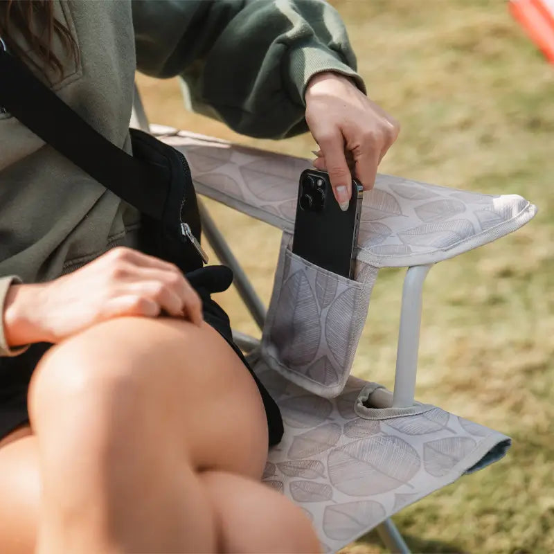 Woman places her phone into the arm pocket of the Soft Neutral Leaf Kickback Rocker.