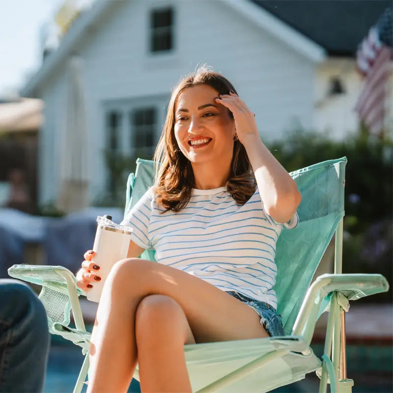 Woman smiles while seated in a pastel green leaf Kickback Rocker beside a pool, holding a tumbler.