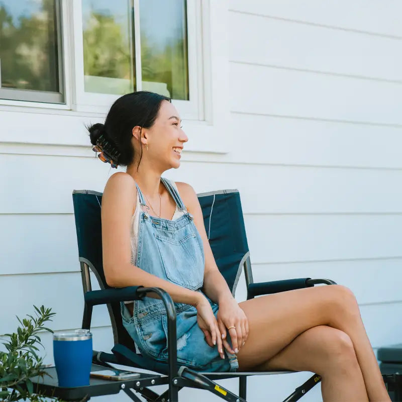 Smiling woman seated outdoors in a Slim-Fold Directors Chair against a white house.