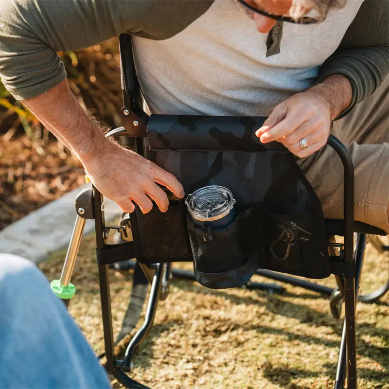 Man sitting in a stealth camo Stowaway Rocker, placing items into the chair's storage pocket.