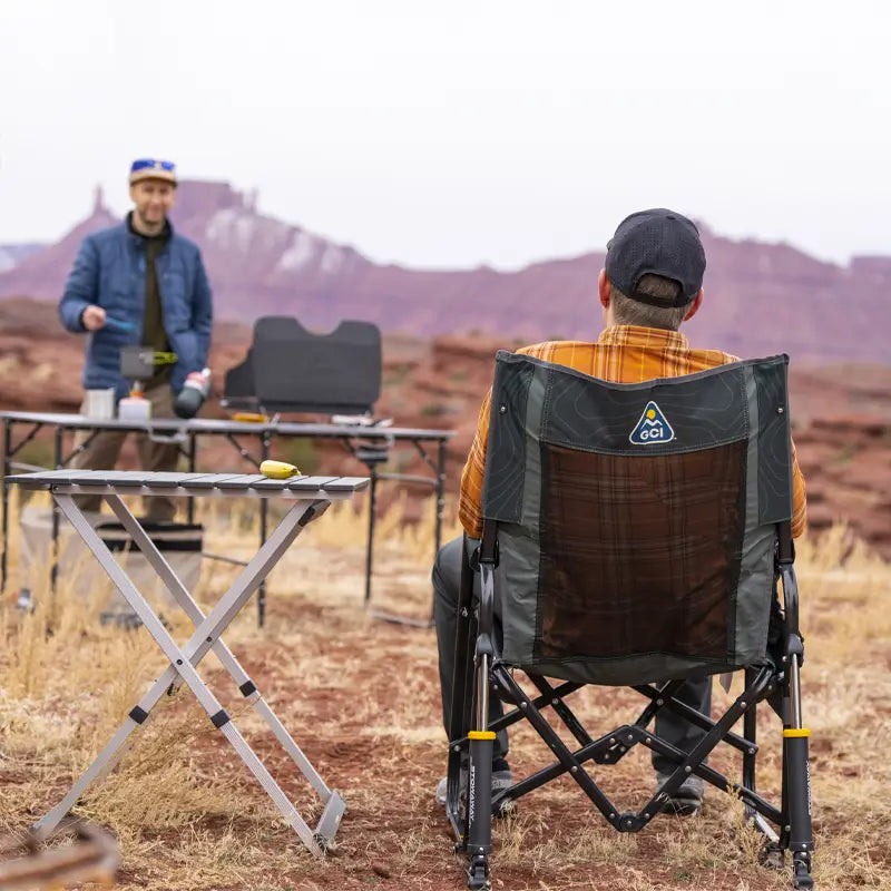 Man sitting in a hunter topo Stowaway Rocker while another cooks at a camp setup in a desert landscape.