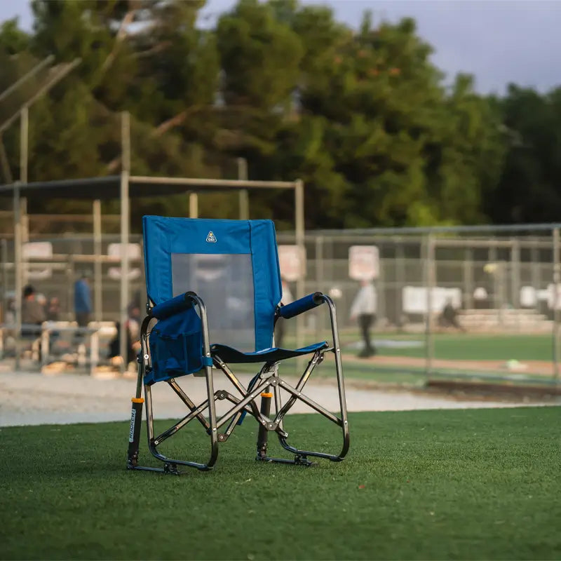 Soft navy Stowaway Rocker sitting empty on a sports field, with fencing and players blurred in the background.