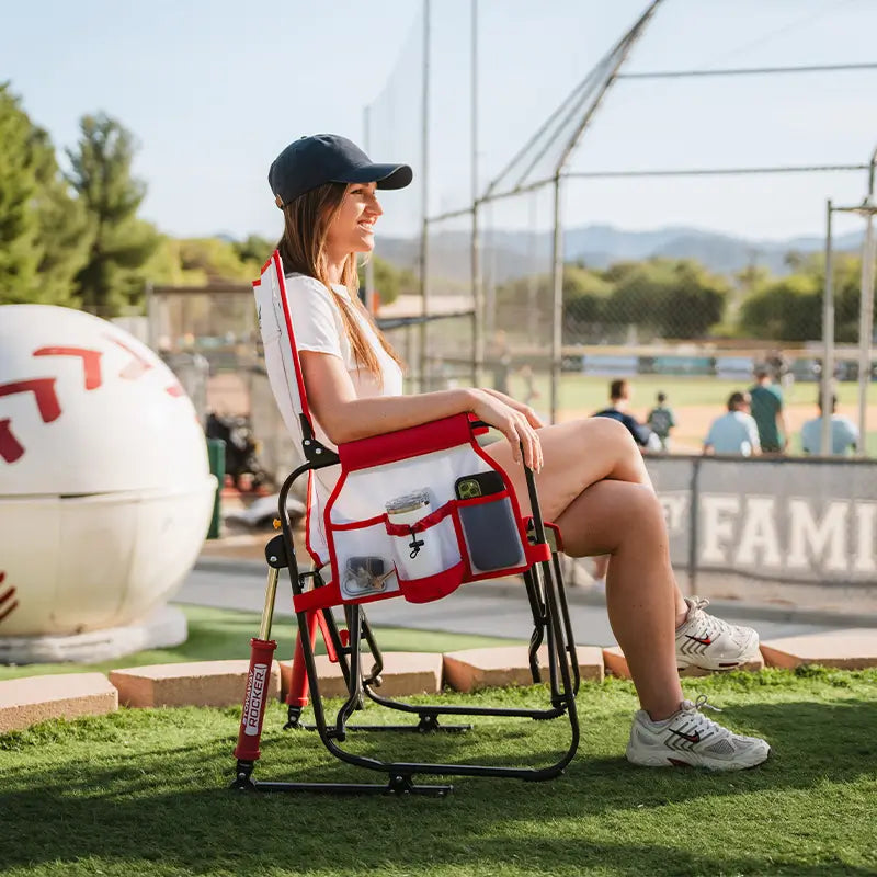 Person sitting on a folding chair with a cup holder and phone holder at a sports field.