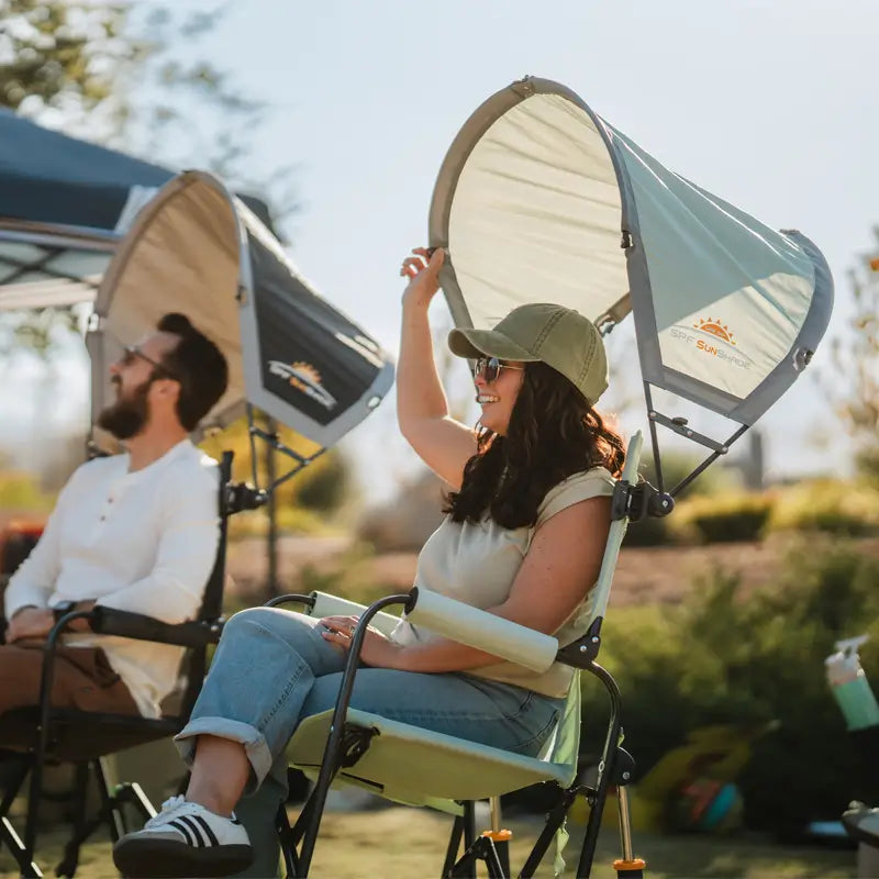 Woman in sunglasses raises a pastel green SPF sunshade while relaxing in a chair at an outdoor event.