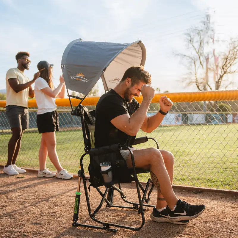Man cheers enthusiastically from a shaded chair with mercury sunshade accessory at a baseball field.