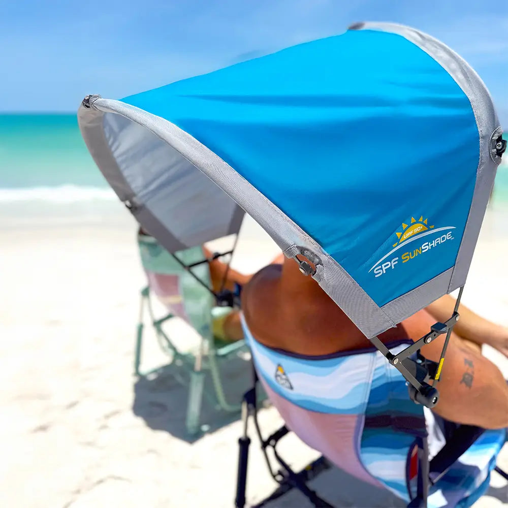 A man sitting in a beach rocker on the beach with the saybrook blue sunshade accessory attached to the chair.