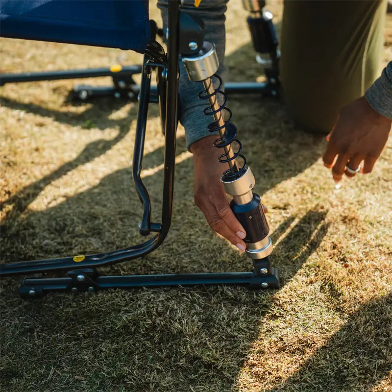 Close-up of a person adjusting one of the shock-absorbing legs of the Adjustable Rocker on dry grass.