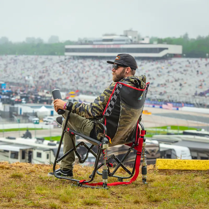 Man in camo jacket lounges in a red-trimmed Adjustable Rocker overlooking a racetrack filled with spectators.