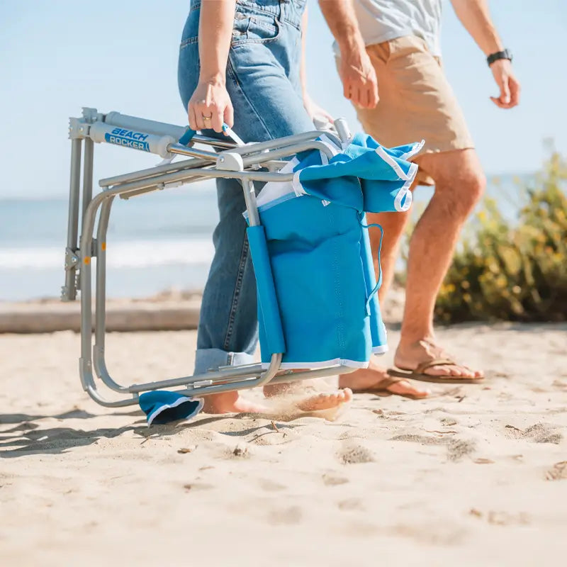 Person walking barefoot on the beach carrying a folded Saybrook Blue Beach Rocker with built-in carry handle.