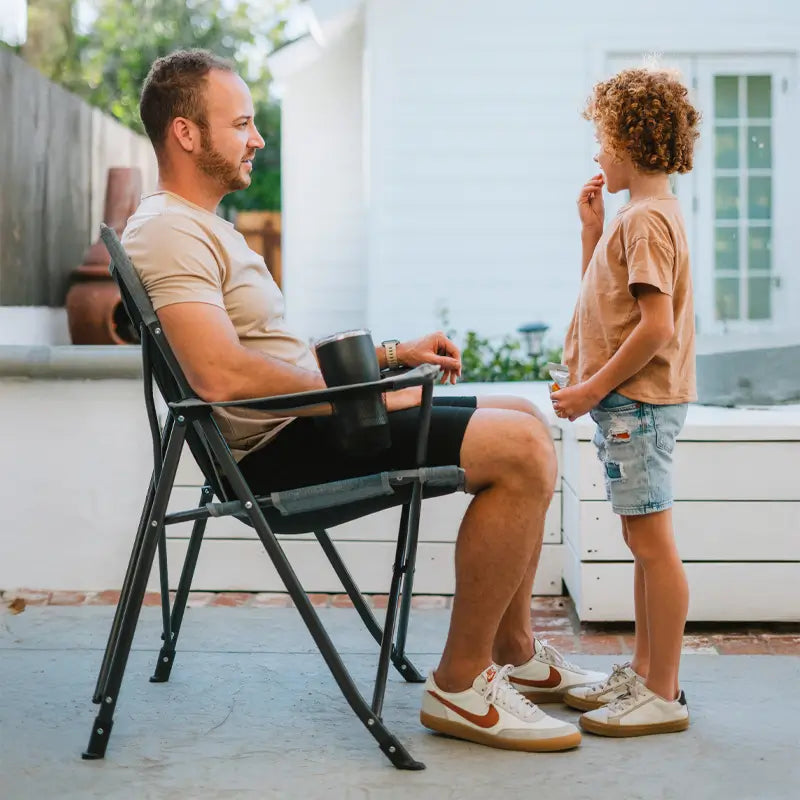 Side view of a man seated in a Comfort Pro Chair, talking to a child in a sunny backyard.