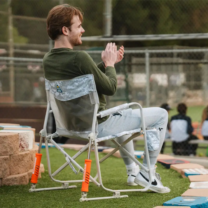 Man claps while seated in a Snow Camo Freestyle Rocker Elite chair at a sports field.
