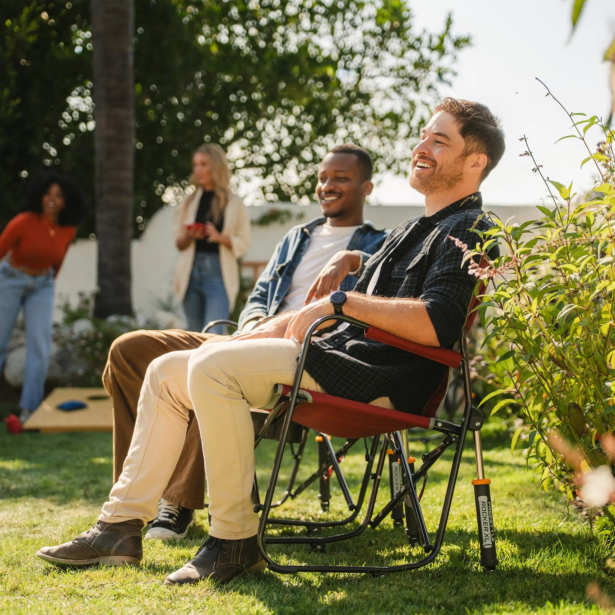 Two men watching people play cornhole while sitting in their freestyle rocker with side table chairs. 