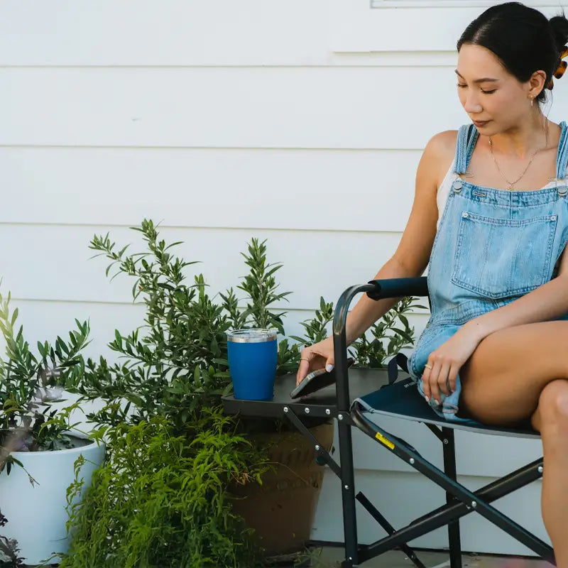 Woman in denim overalls relaxing on a patio in a Slim-Fold Directors Chair with side table and drink.