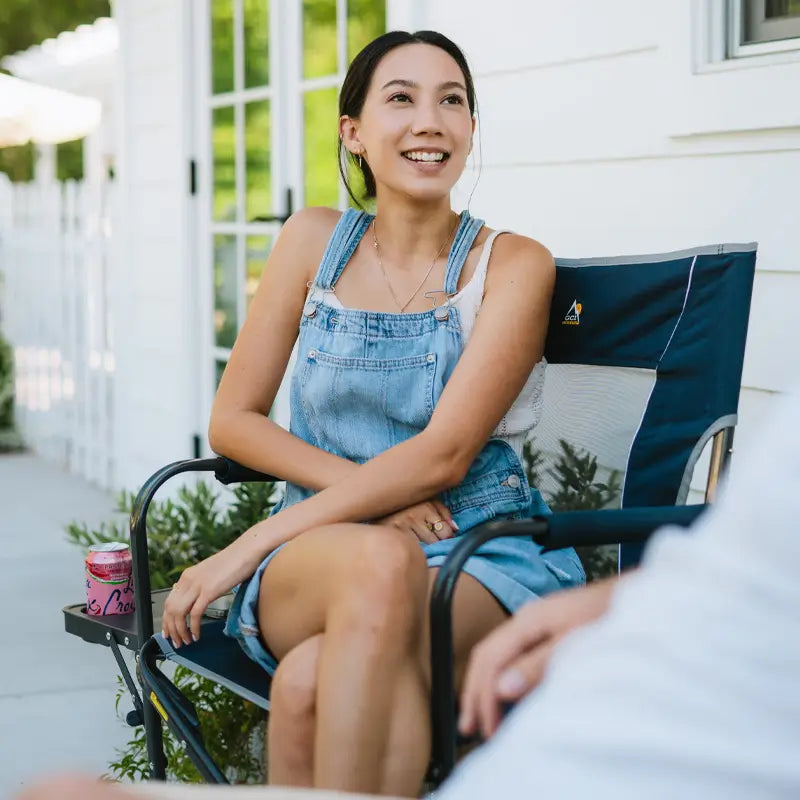Woman chatting on a patio while seated in the navy Slim-Fold Directors Chair with built-in side table.