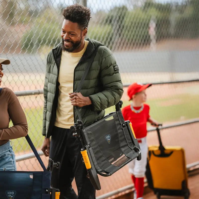 Man carrying the folded dark charcoal Stadium Rock-Cliner with shoulder strap at a baseball field.