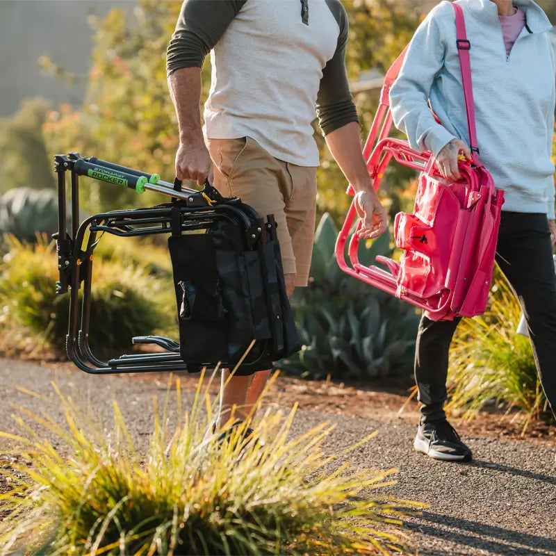 Two people walking outdoors carrying folded Stowaway Rockers, one in stealth camo and one in bright pink.