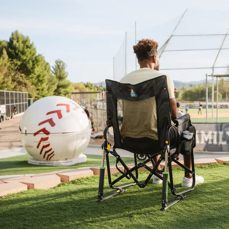 Man seated in the stealth camo Stowaway Rocker, overlooking a baseball field near a large baseball sculpture.
