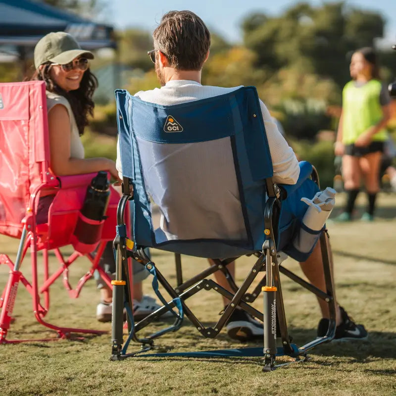 Man sitting in a soft navy Stowaway Rocker at a sports field, talking with a woman in a pink chair beside him.