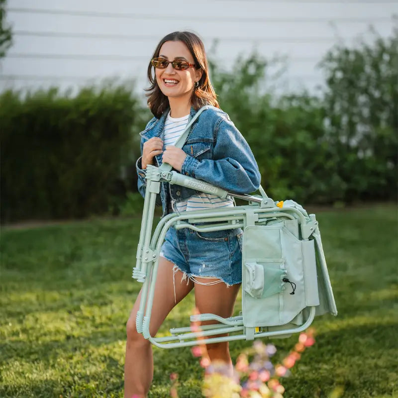 Woman carrying a folded pastel green Stowaway Rocker over her shoulder in a grassy yard.