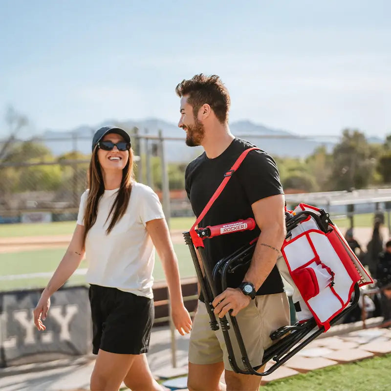 Two people walking outdoors with a red and white chair on a sunny day.