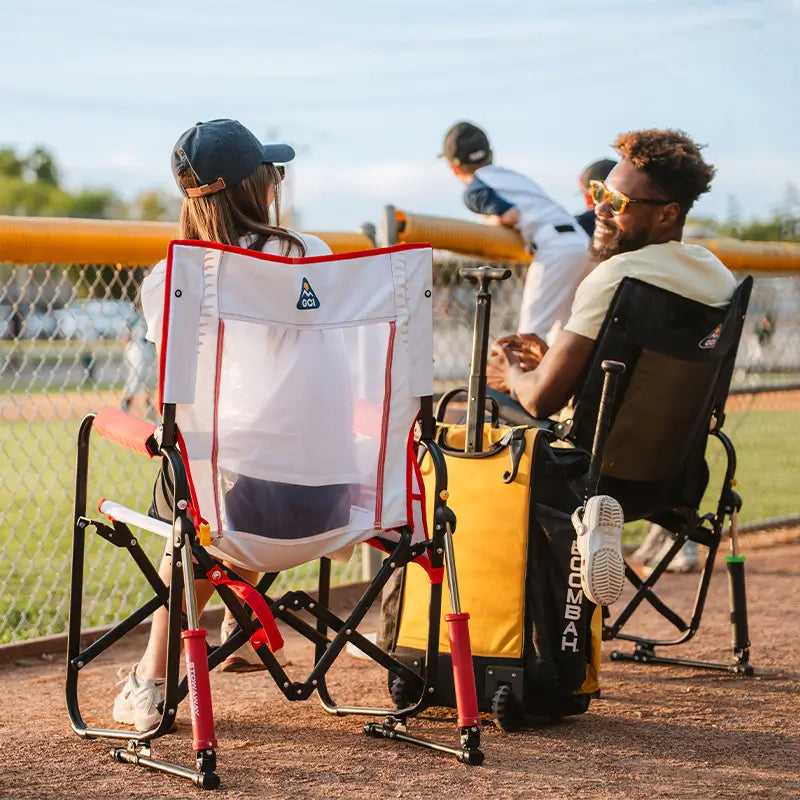 Two people sitting on folding chairs at a baseball game with sports equipment.