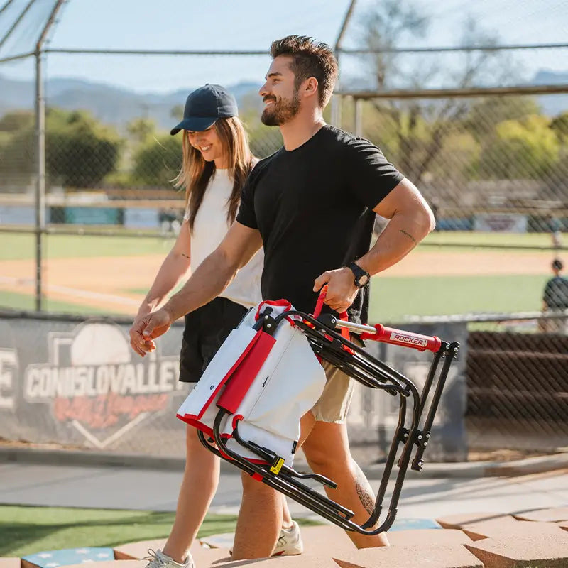 Two people walking with a portable folding chair on a sports field.