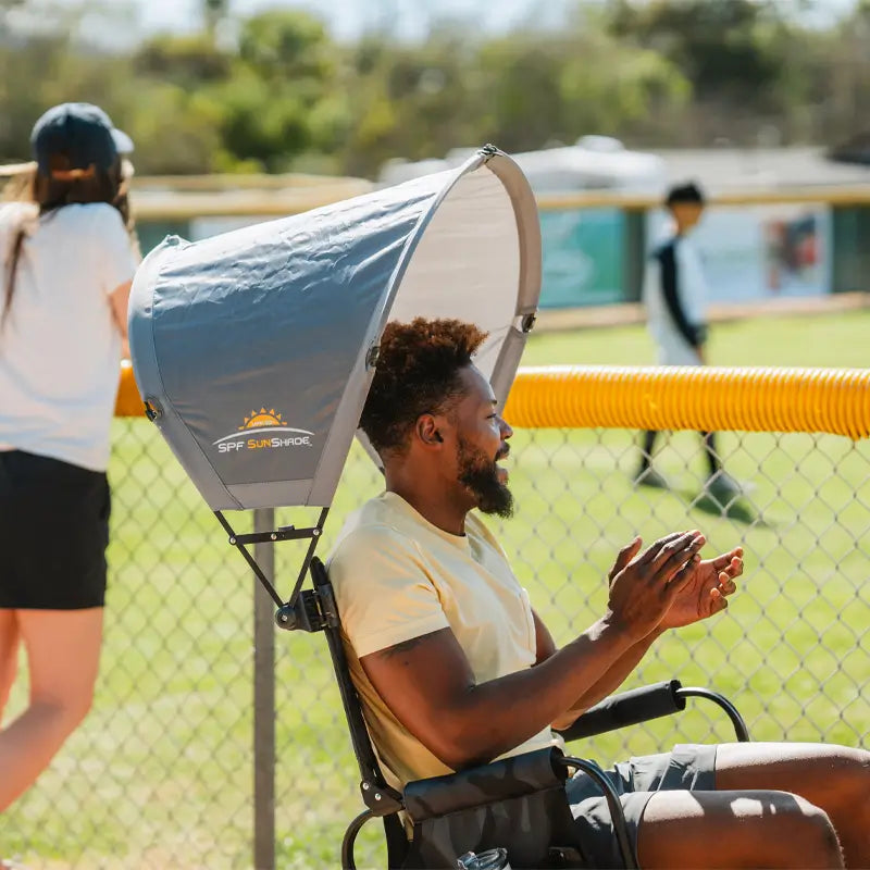 Man sits and claps while shaded by mercury sunshade accessory at a baseball field.