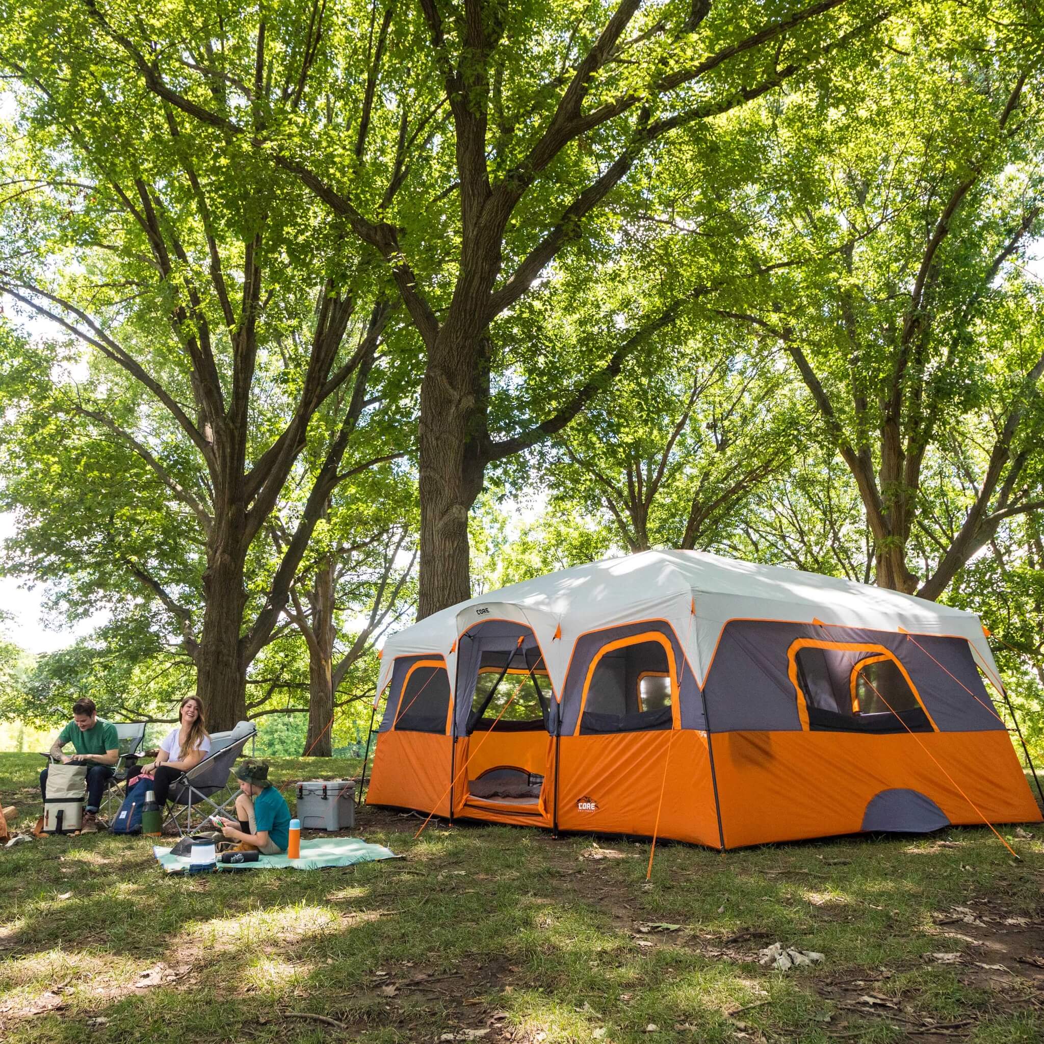 CORE 12 person straight wall cabin tent in orange and gray set up at a campsite with campers relaxing under trees beside the tent.