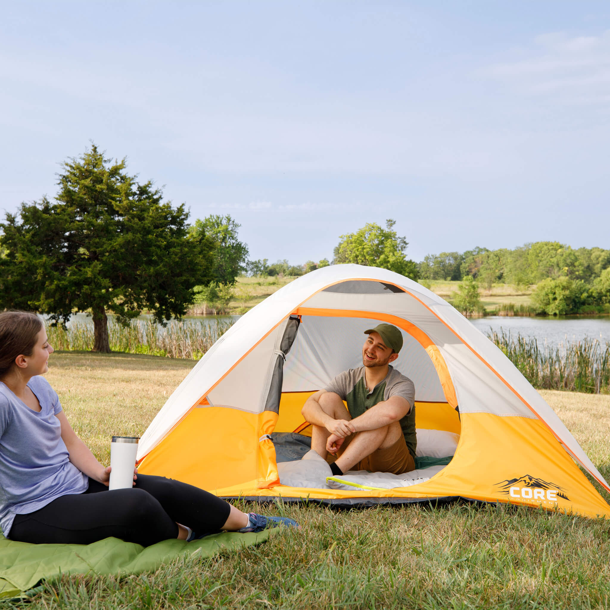 Two campers sitting by CORE 3 person dome tent near a lake, with open door showing spacious interior for outdoor comfort.