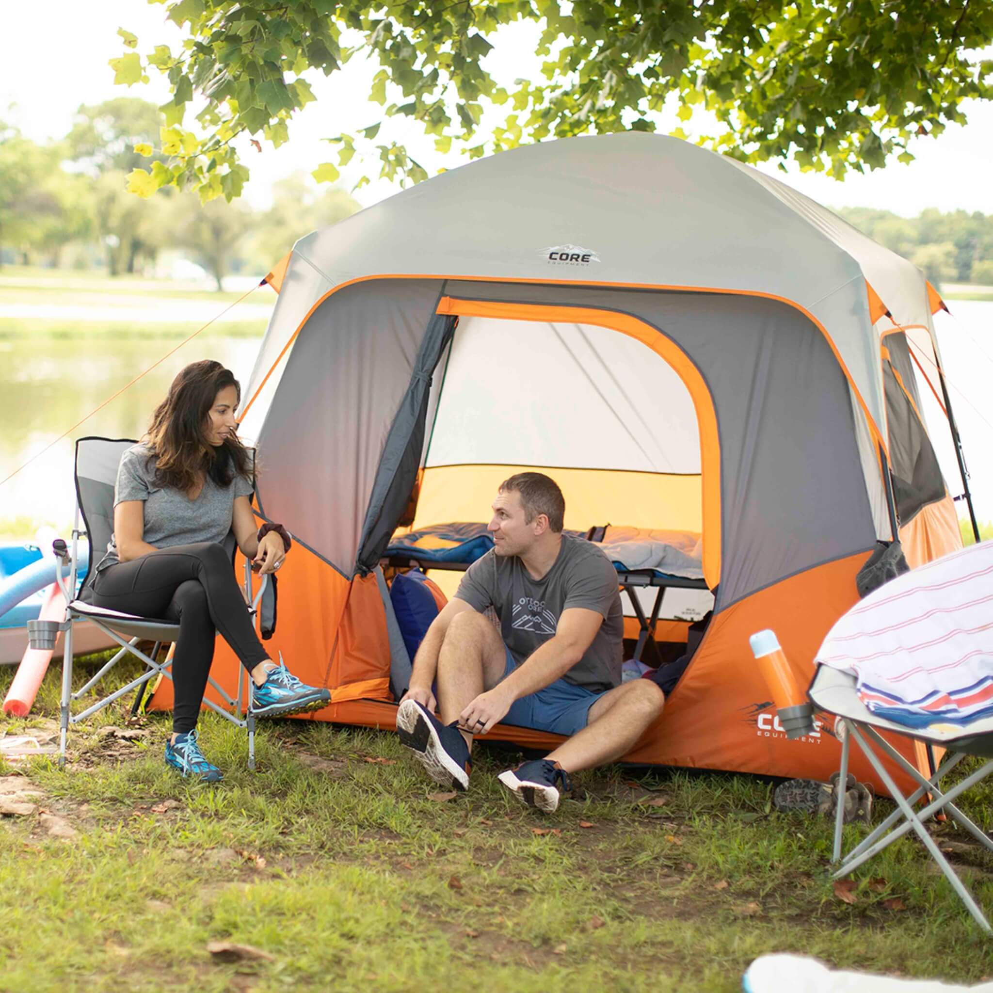 Two people sitting on camping chairs outside a CORE 4 person straight wall cabin tent in orange and gray. They are near trees with a lake in the background. One person is tying their shoes while the other is holding a water bottle.