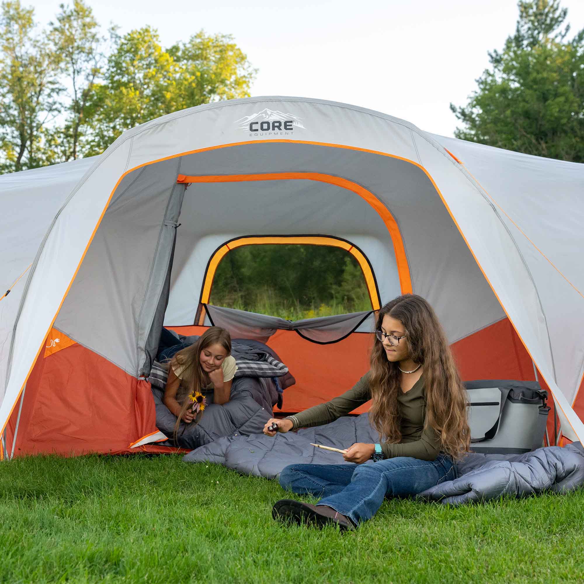 Two people sitting outside a CORE 9 person dome tent in a grassy area with trees in the background.